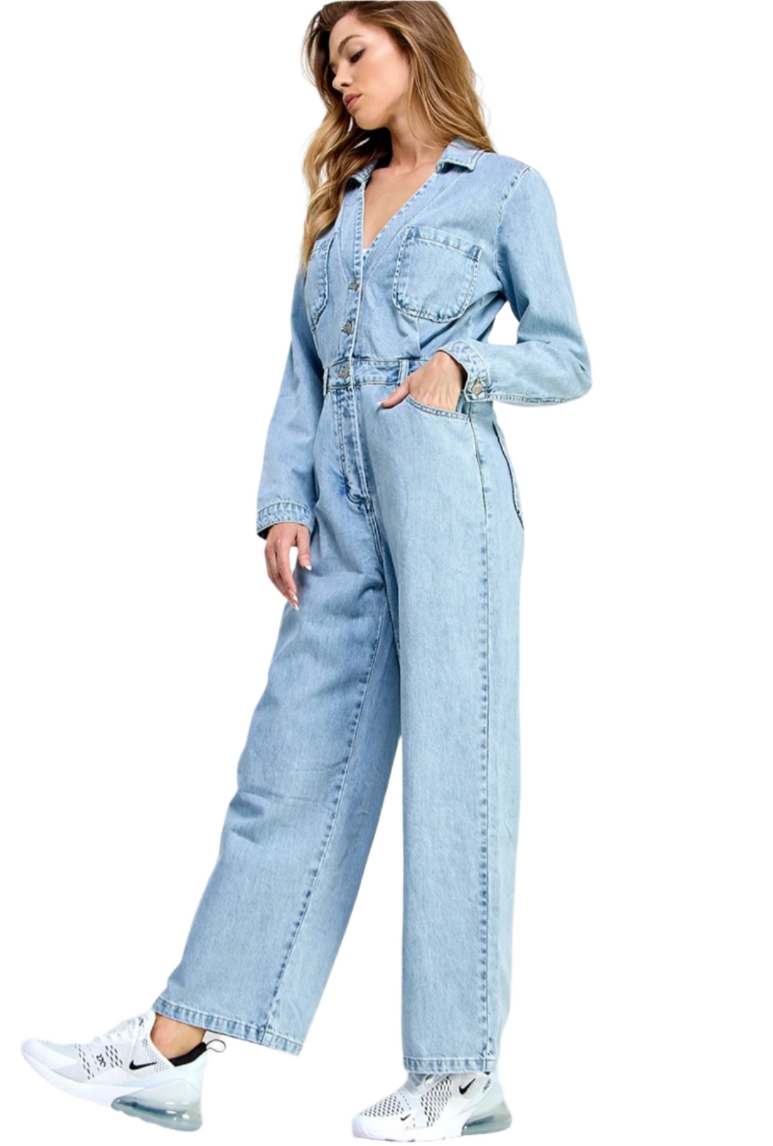 A female model wearing a blue denim jumpsuit with a collar, long sleeves, and straight pants, paired with white sneakers.