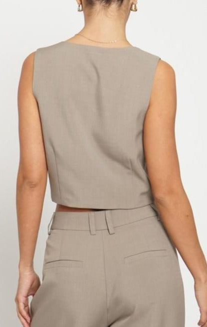 A female model wearing a beige woven vest with a deep V-neckline, two-button closure, and a pocket detail.