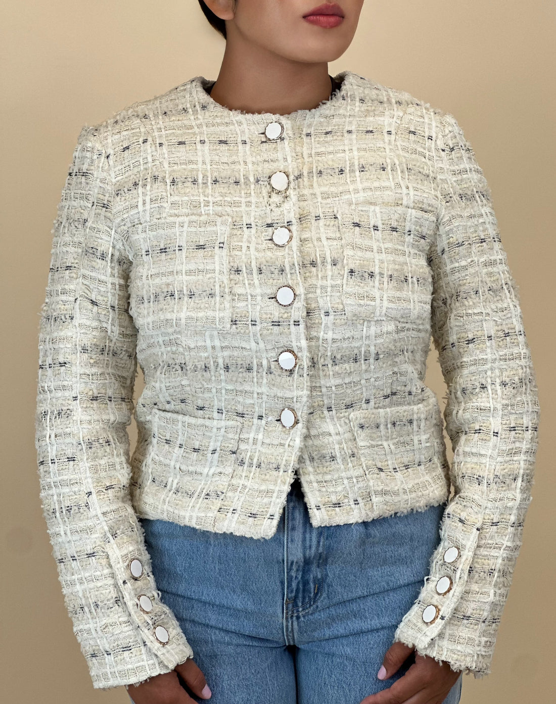 Person wearing a textured beige jacket over a dark top and blue jeans on a beige background