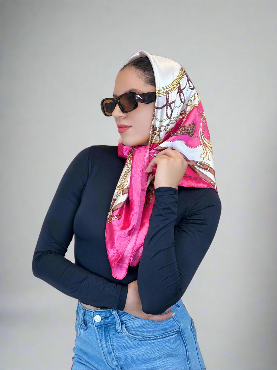 Woman wearing a pink and gold patterned scarf, sunglasses, and a navy long-sleeve top with a beige background.