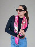 Woman wearing a pink scarf with a pattern, black sunglasses, and blue jeans against a beige background