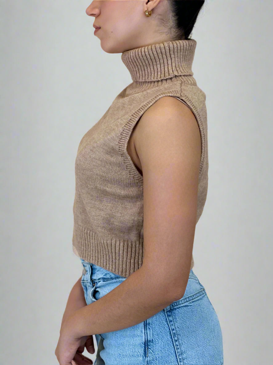 Person wearing a beige sleeveless knitted top and blue jeans against a plain background