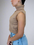 Person wearing a beige sleeveless knitted top and blue jeans against a plain background