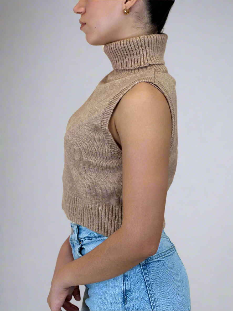 Person wearing a beige sleeveless knitted top and blue jeans against a plain background