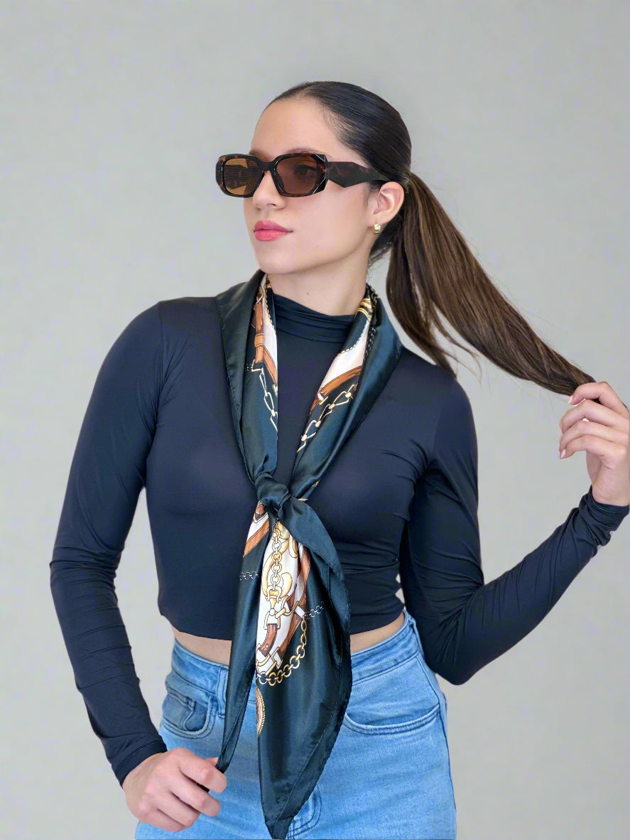 Woman wearing a black long-sleeve top, blue jeans, and sunglasses with a patterned scarf around her neck against a plain background.