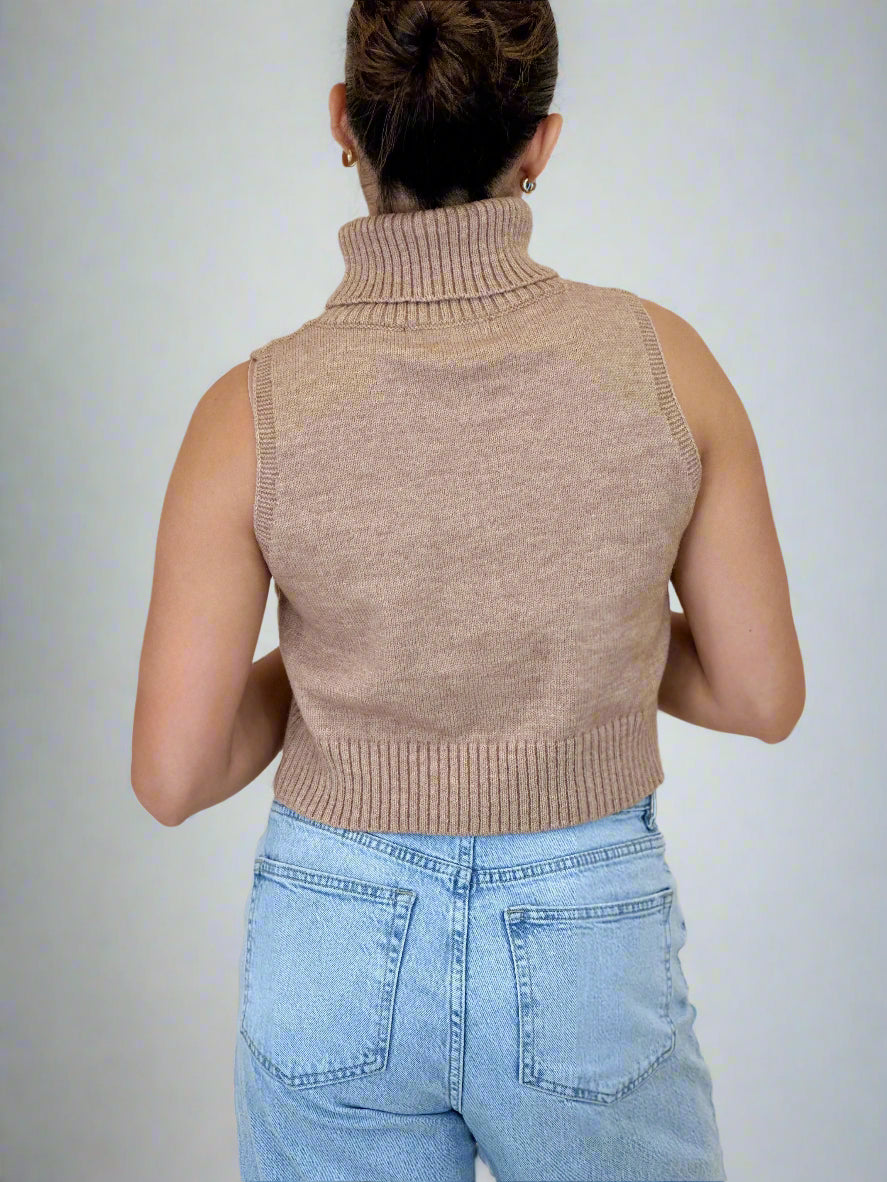 Person wearing a beige sleeveless knitted top and light blue jeans on a beige background