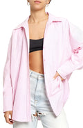 A woman wearing a pink oversized button down shirt with collared neckline and long sleeves, paired with blue jeans. She is also wearing a black sports bra and a necklace.