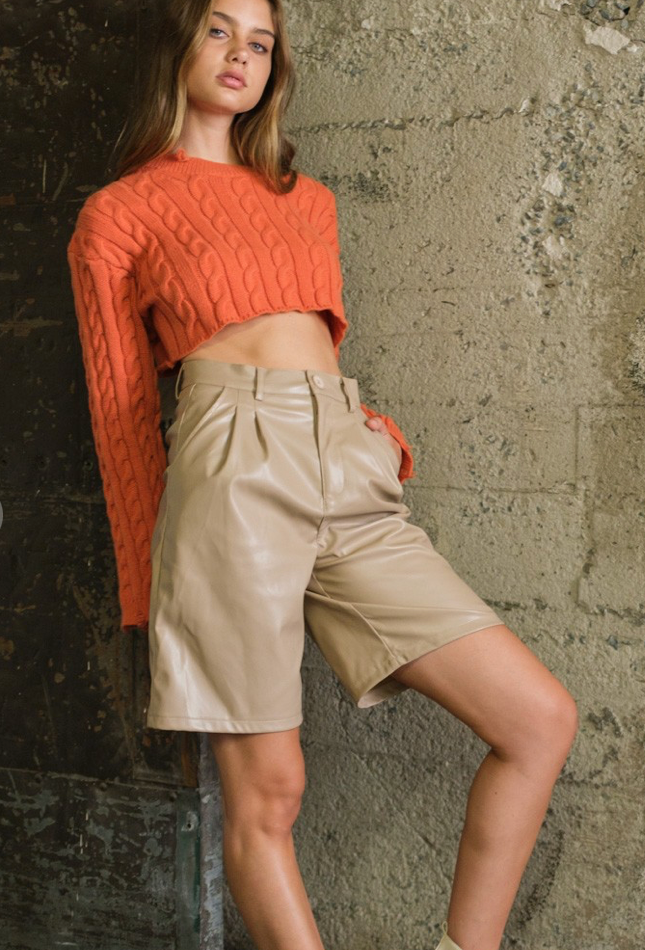 A model wearing high-waisted beige faux leather Bermuda shorts with a pleat detail and a knit orange top.