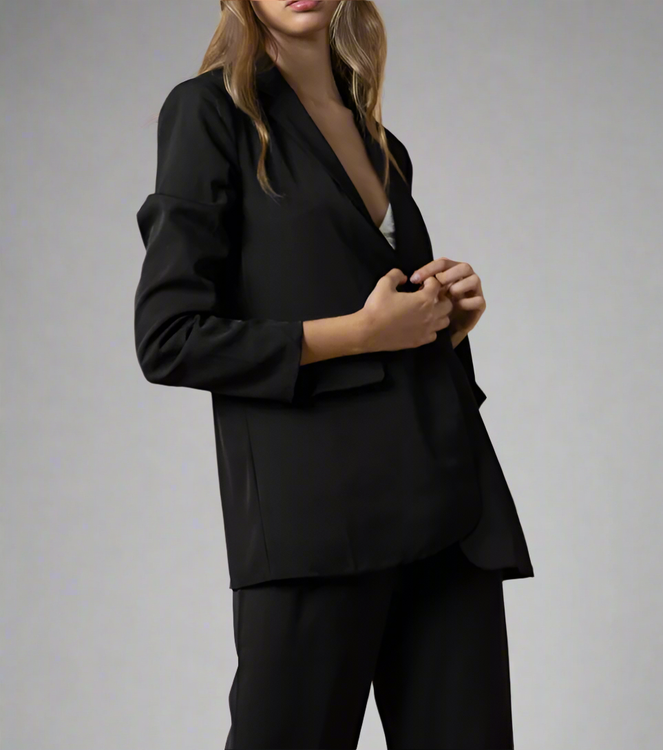 A female model wearing a black oversized single-breasted blazer with buttoned front and flap pockets on the front, paired with black trousers.