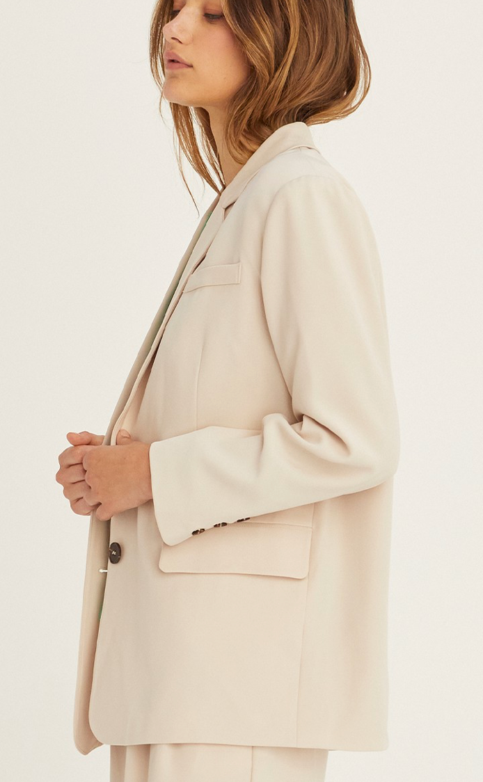 A woman wearing a beige single-breasted blazer with a notched collar and a front flap pocket, paired with a green top and trousers.